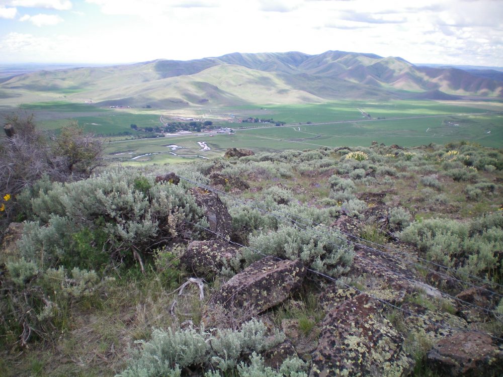 The summit boulders and scrub atop Peak 6145 with fence posts and barbed wire on the ground nearby. The hamlet of Picabo, ID is on the valley floor. The Picabo Hills are in the distance. Livingston Doug Photo
